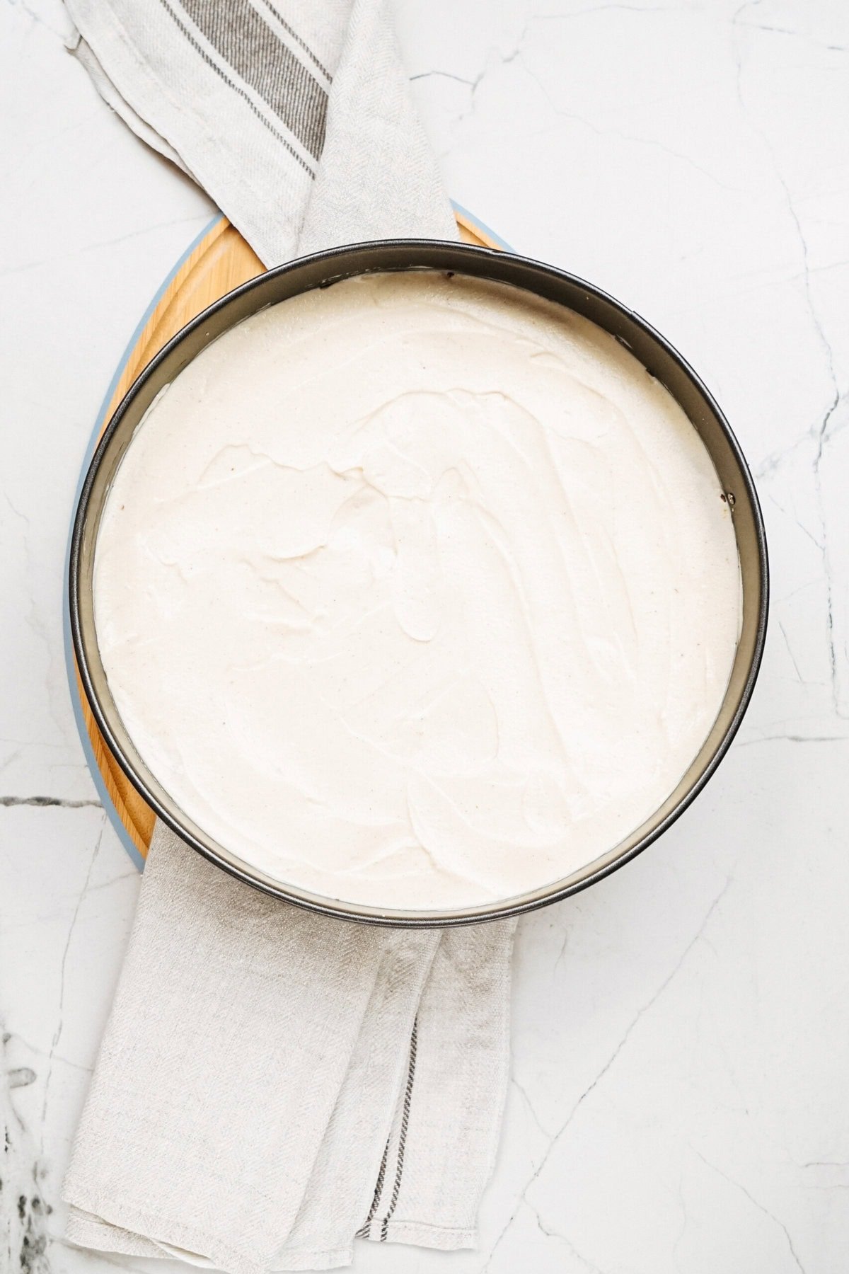 A round cake pan filled with creamy white batter rests on a gray-striped cloth on a marble surface.