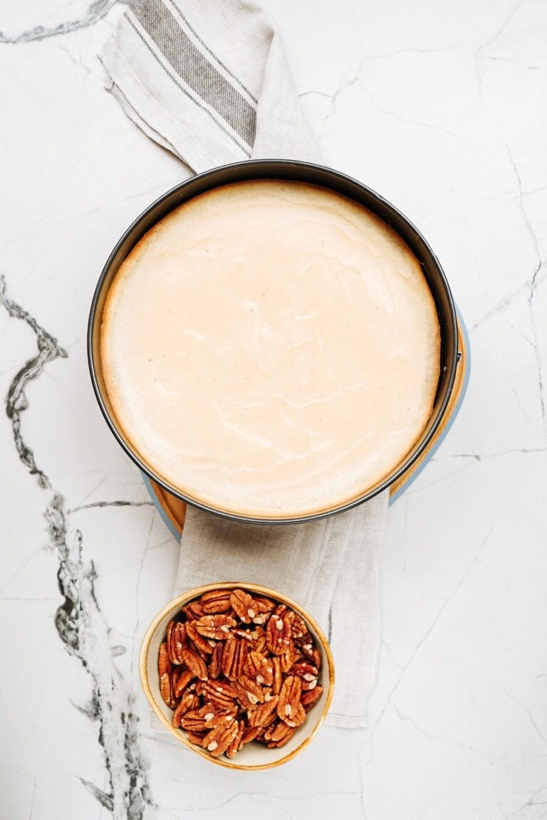 A round cheesecake sits on a marble surface alongside a bowl of pecans.