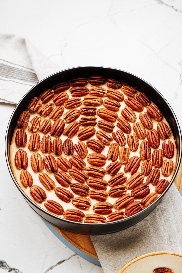 Pecan-topped cheesecake in a round black pan on a marble surface.