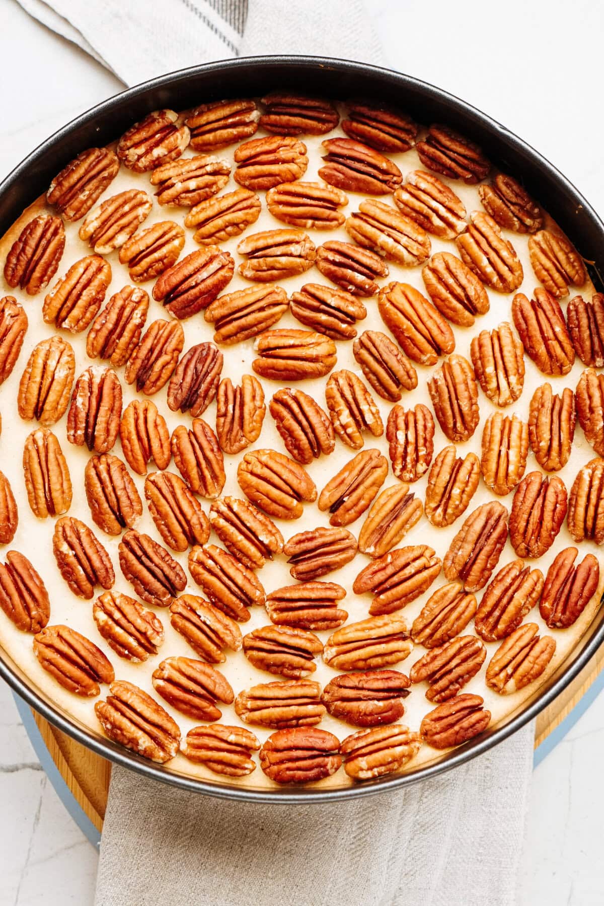 A round pecan pie with a crusted edge, topped with whole pecans, sits on a light-colored towel on a wooden surface.