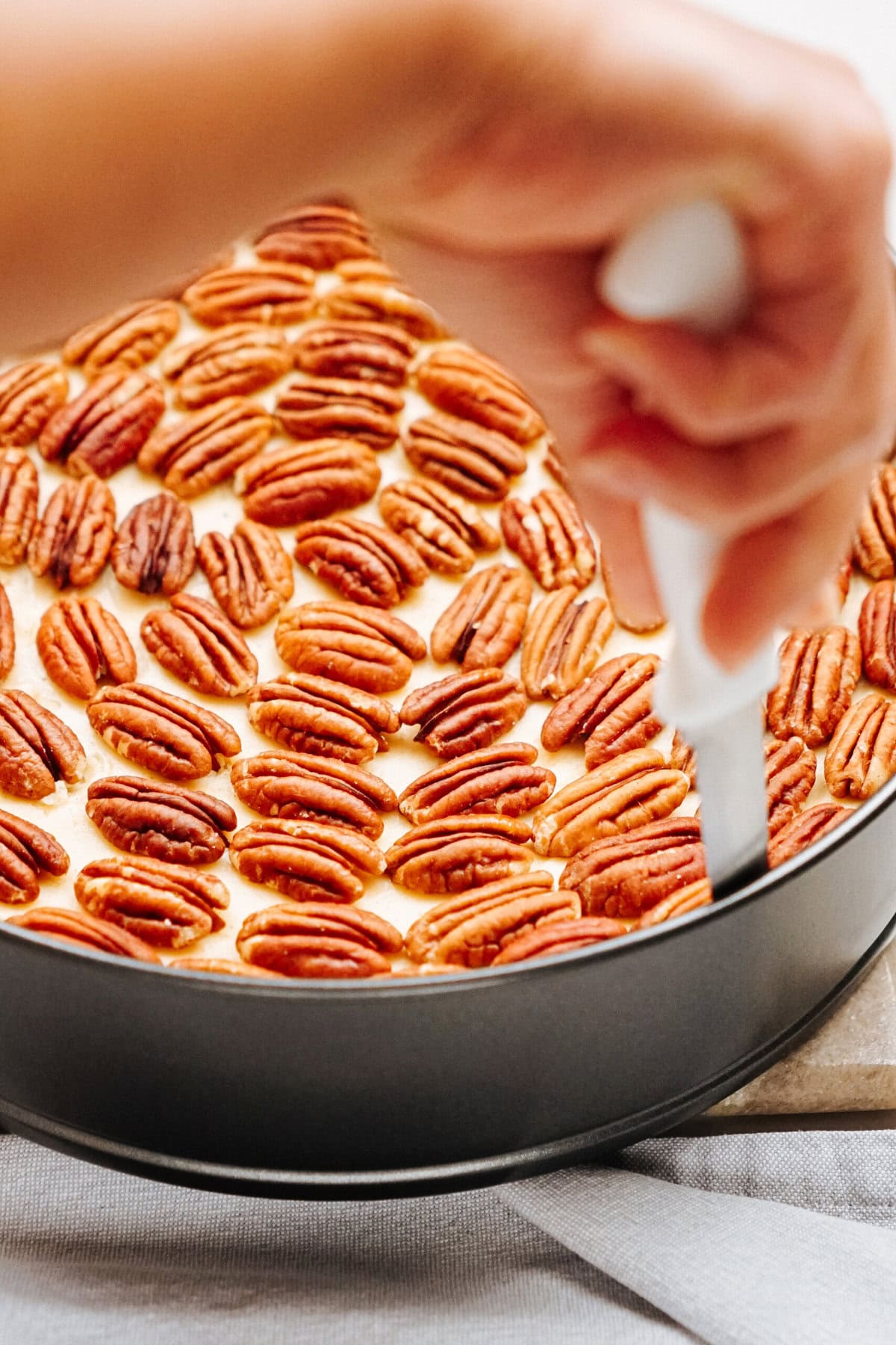 A hand holding a knife is slicing a dessert topped with rows of pecans in a round pan.