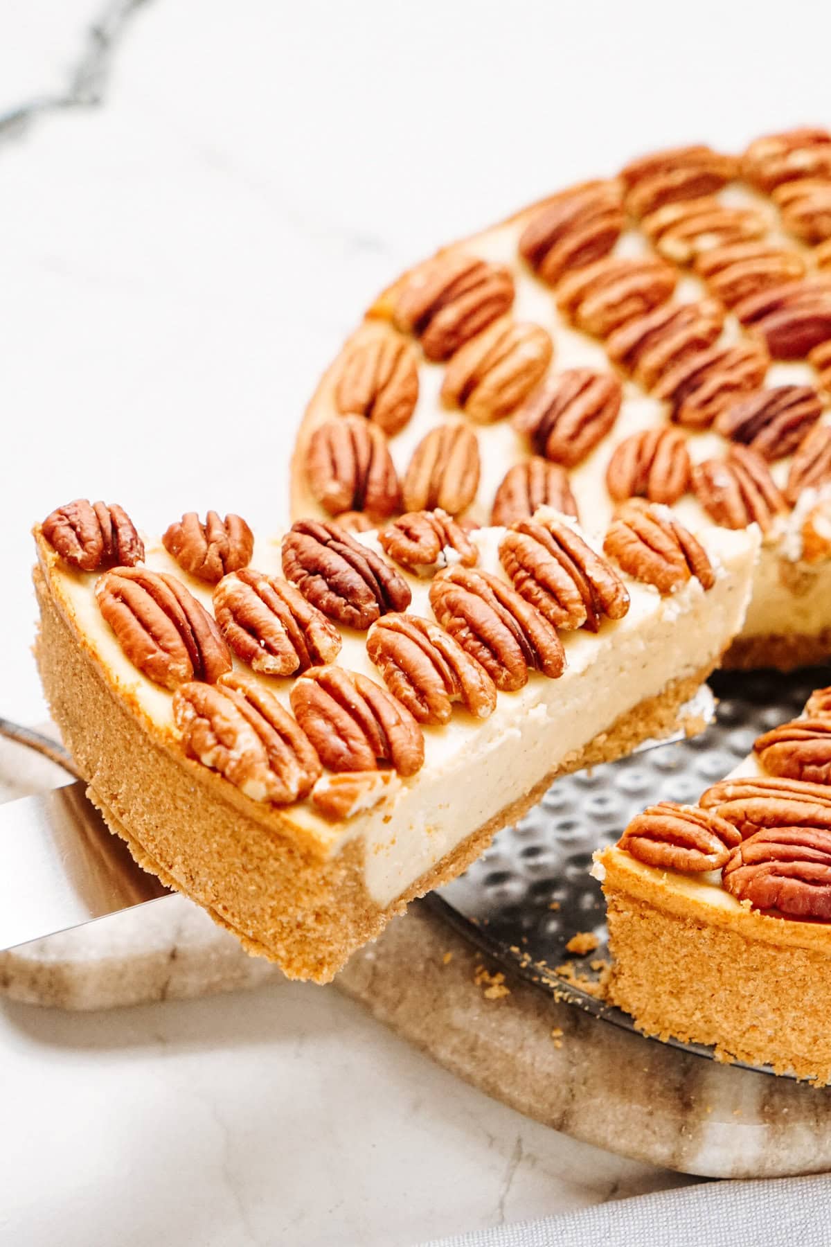 A slice of pecan-topped cheesecake being lifted from a round cake on a table.