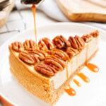 A slice of cheesecake topped with pecans is drizzled with caramel sauce on a white plate.