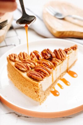 A slice of cheesecake topped with pecans is drizzled with caramel sauce on a white plate.