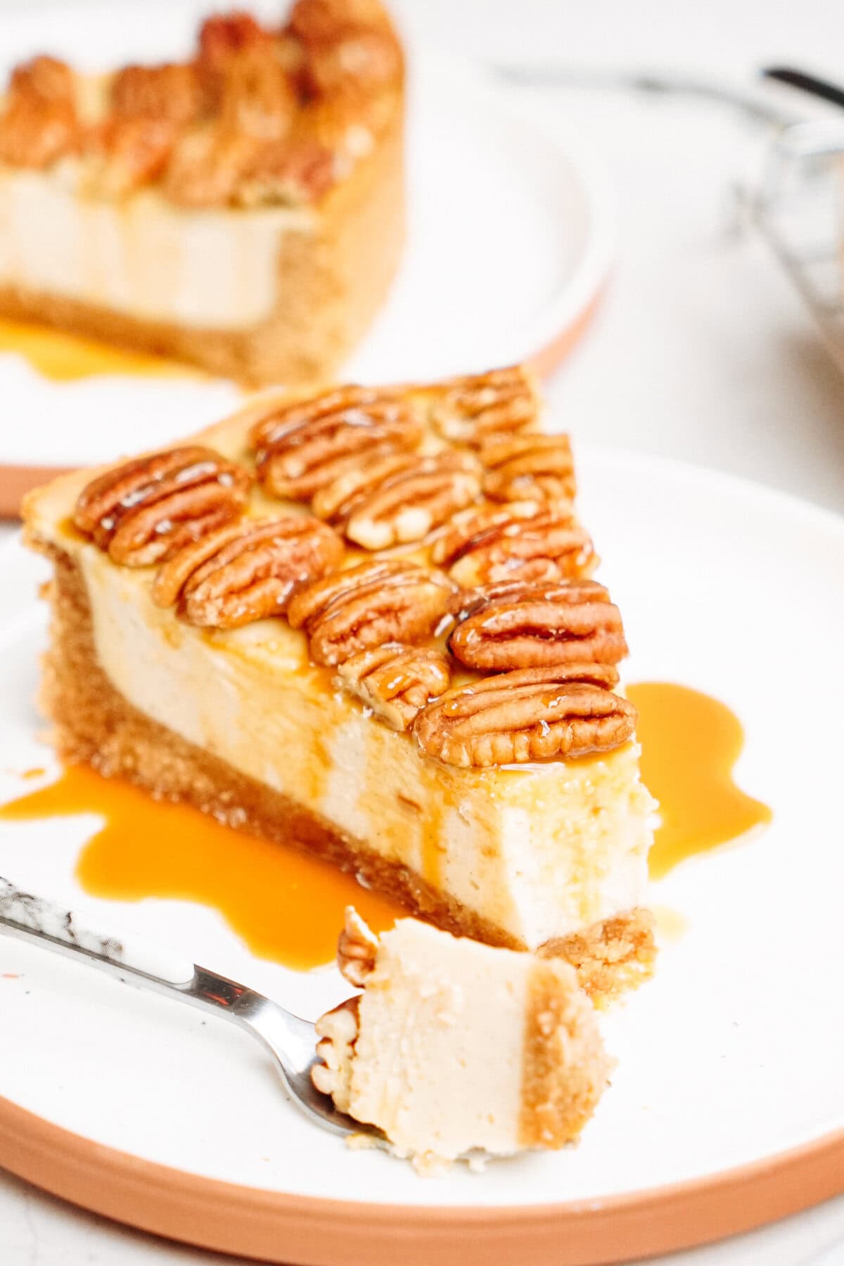 A slice of pecan cheesecake on a white plate, topped with pecans and caramel sauce, with a fork holding a small piece beside it.