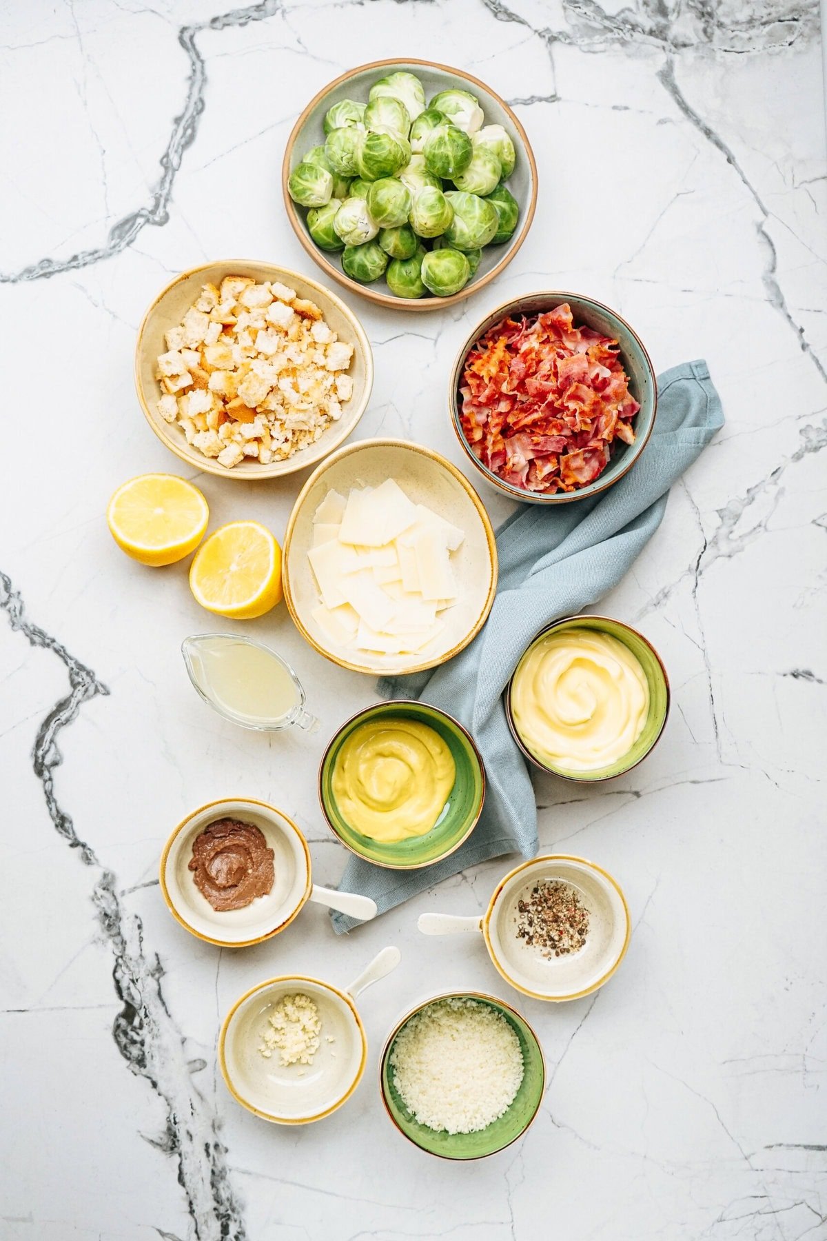 Ingredients for a salad on a marble surface include Brussels sprouts, bacon bits, croutons, lemon halves, dressing, cheese, and seasonings in separate bowls.