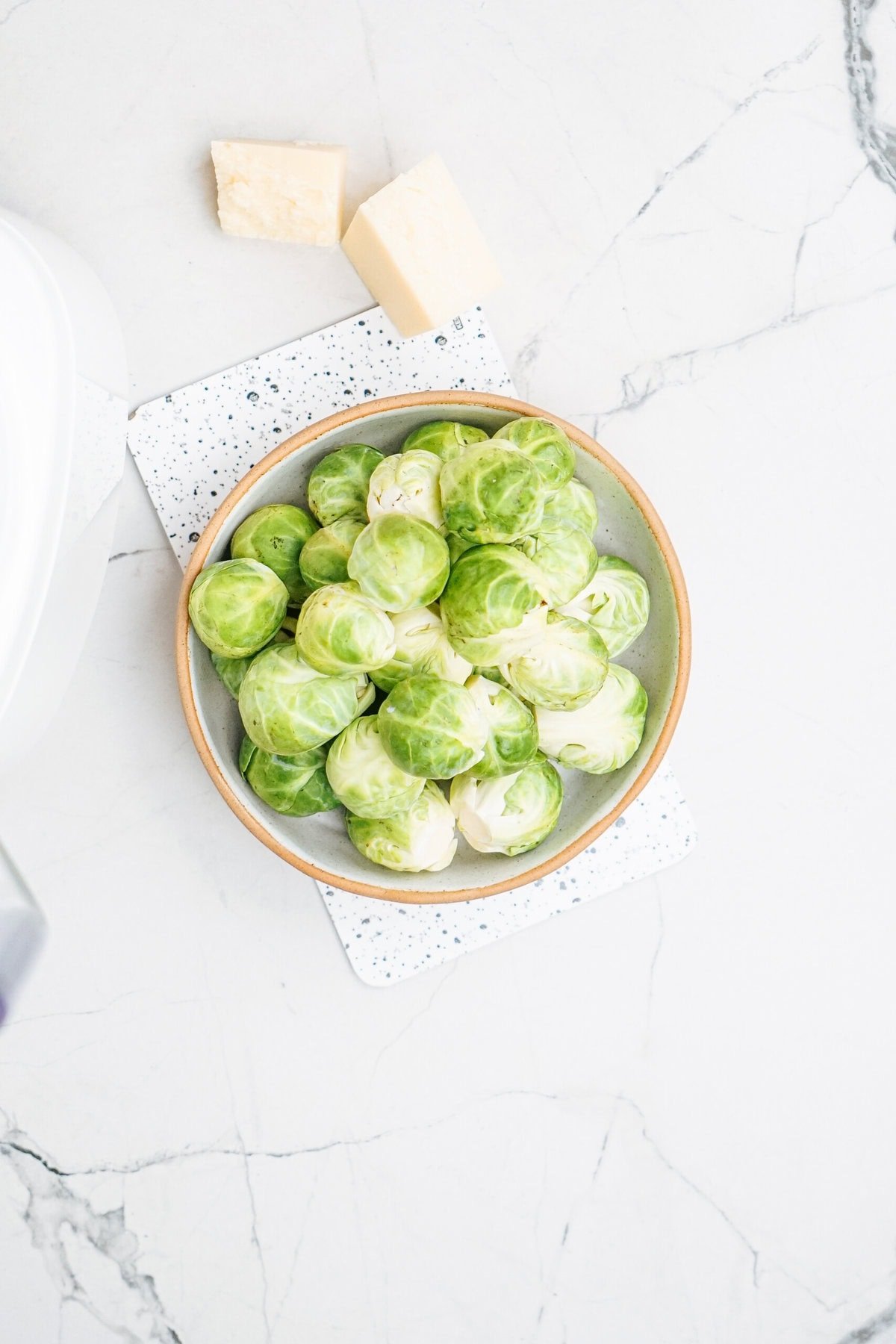 A bowl of Brussels sprouts on a marble surface with two small blocks of cheese nearby.