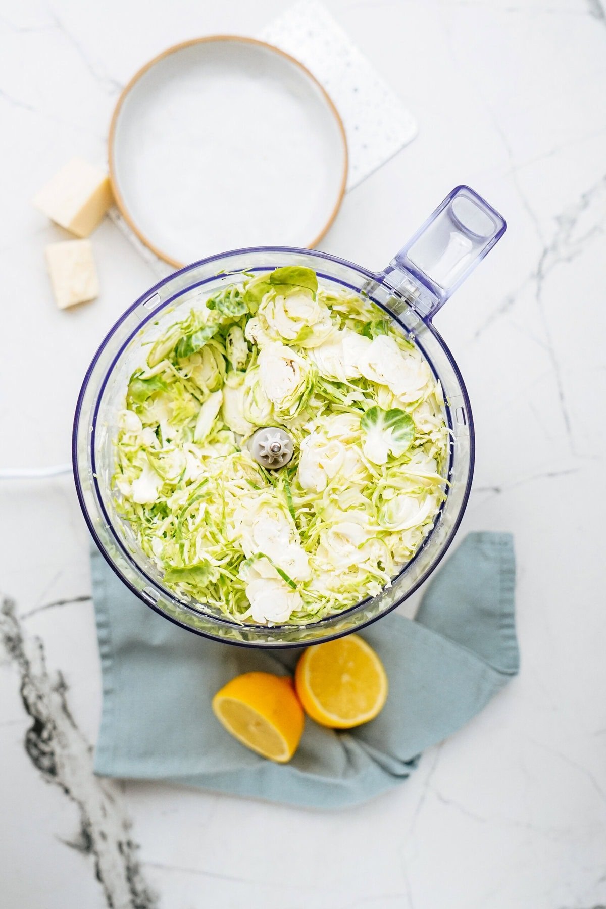 Food processor with sliced Brussels sprouts on a marble countertop, accompanied by two lemon halves and a round dish.