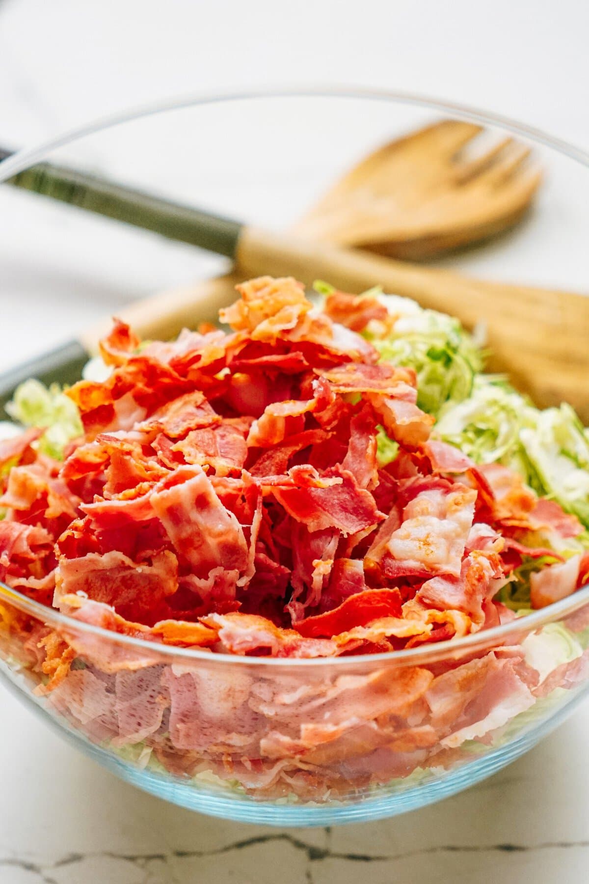 A glass bowl filled with chopped bacon and shredded cabbage, with wooden utensils in the background.