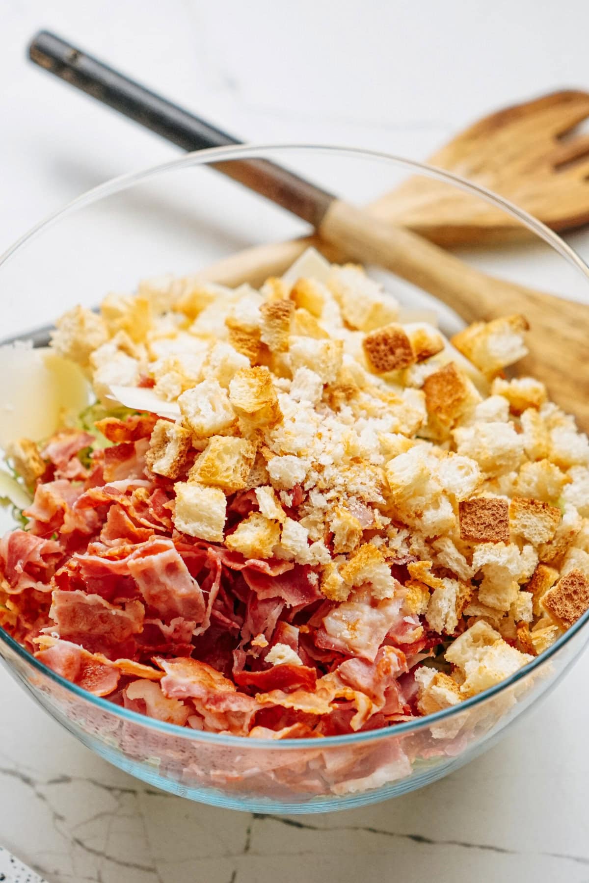 A glass bowl filled with chopped bacon, croutons, and shredded cheese sits on a marble surface. A wooden fork and spoon are in the background.