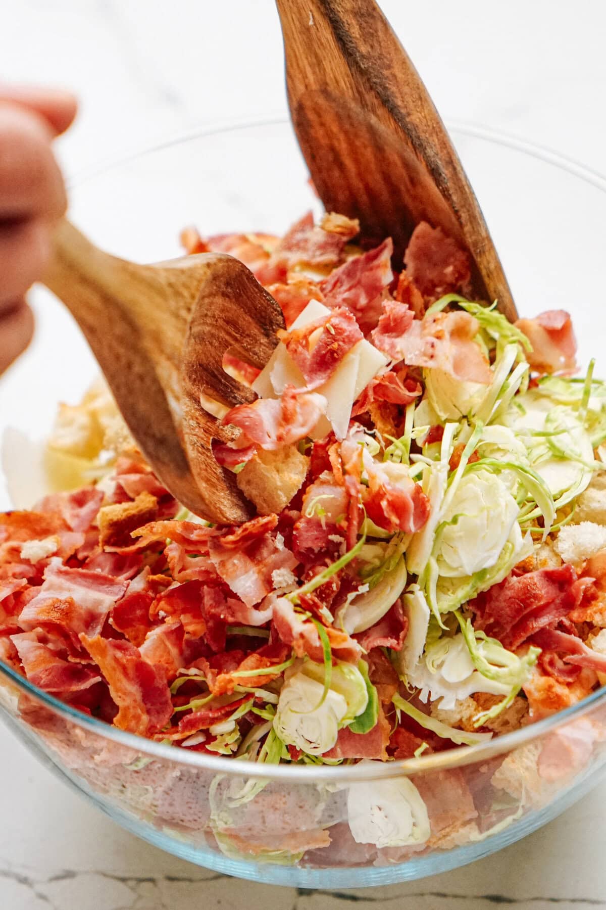 A person tossing a salad with wooden utensils. The salad contains sliced Brussels sprouts, crispy bacon, and shaved Parmesan in a clear glass bowl.