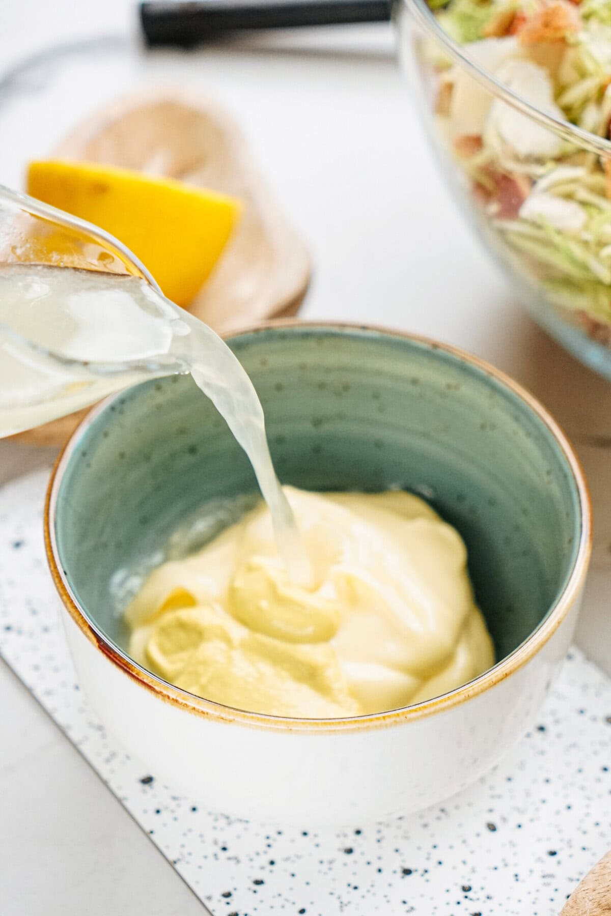 Lemon juice being poured into a bowl of mayonnaise on a speckled cutting board, next to a lemon wedge and a salad bowl.