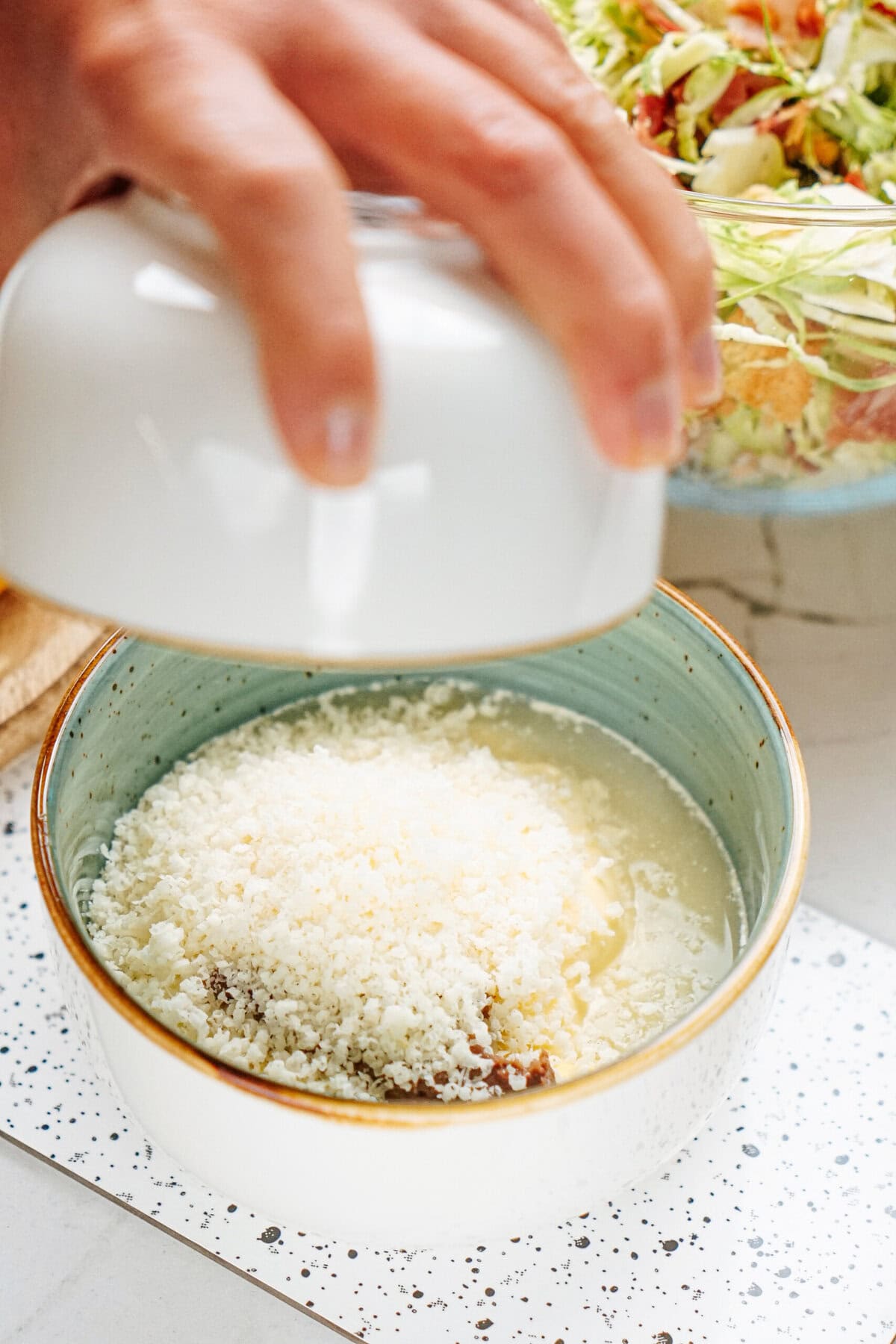 A hand holding a lid over a bowl of salad topped with grated cheese, with another bowl of shredded vegetables in the background.