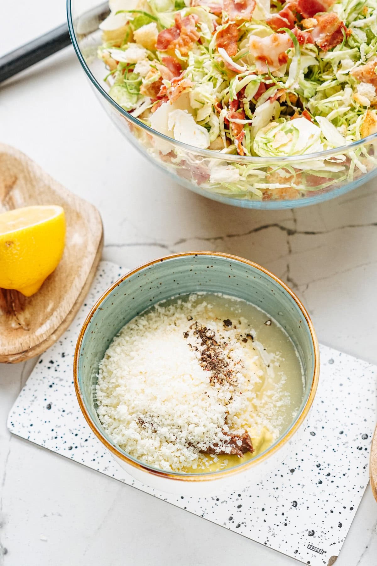 A bowl of grated cheese, anchovies, and pepper is prepared next to a lemon wedge and a bowl of salad with lettuce, bacon, and croutons on a marble countertop.