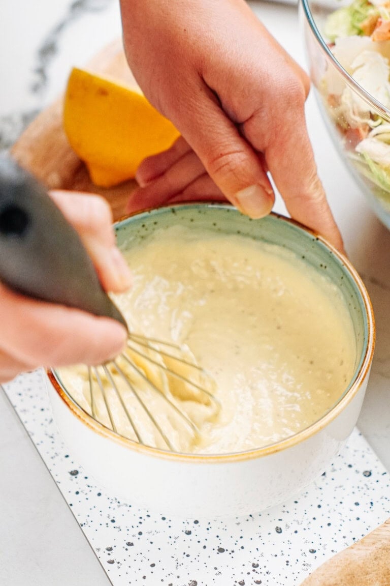 Person whisking creamy dressing in a small bowl with a whisk, next to a sliced lemon.