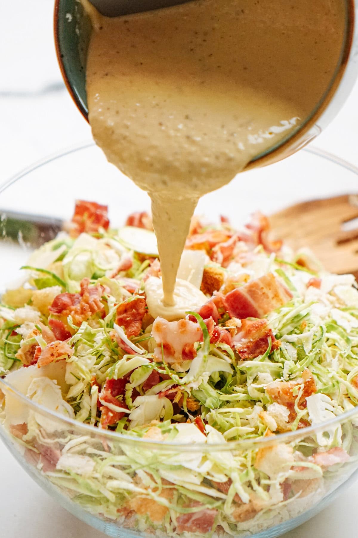 A creamy dressing is being poured over a salad made of shredded cabbage, bacon bits, croutons, and pieces of cheese in a glass bowl, with a wooden spoon in the background.