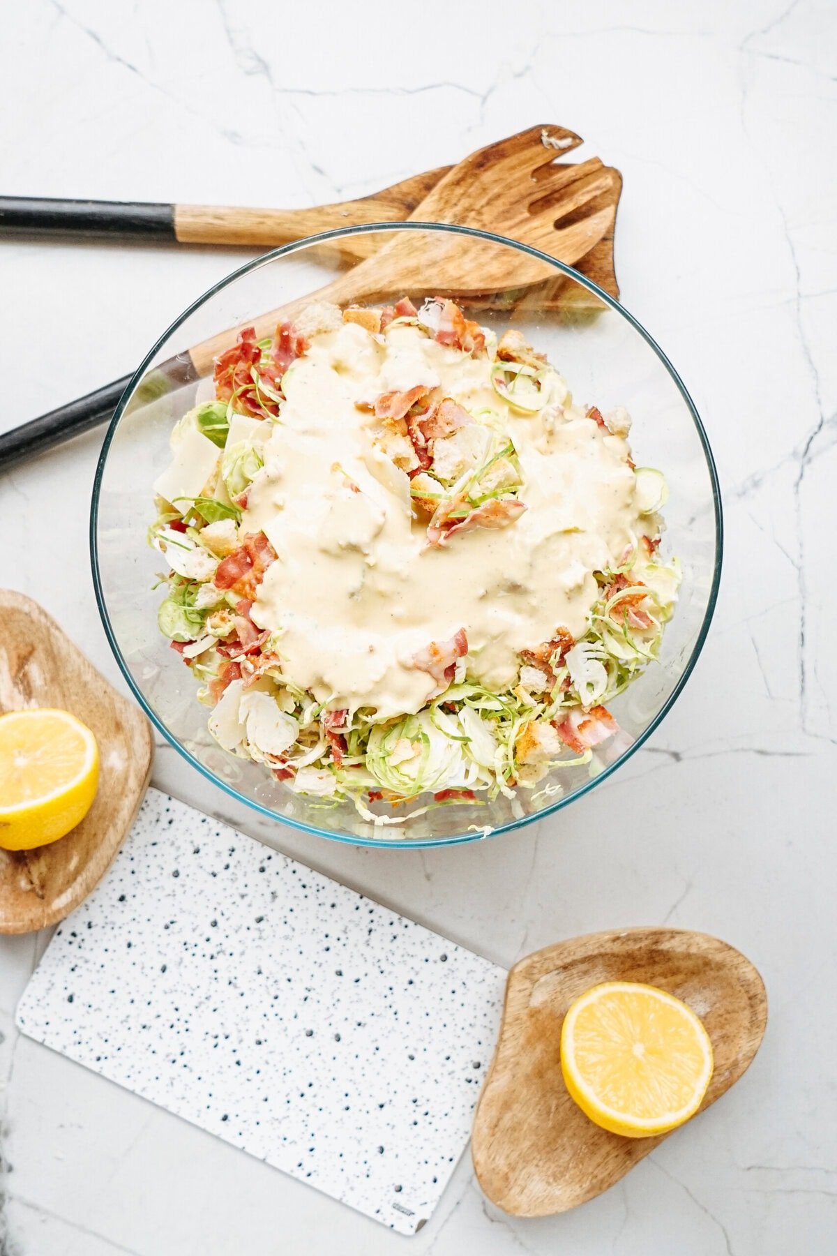 A clear bowl of salad with shredded greens, diced bacon, and creamy dressing on a marble countertop. Wooden salad tongs and sliced lemons are nearby.