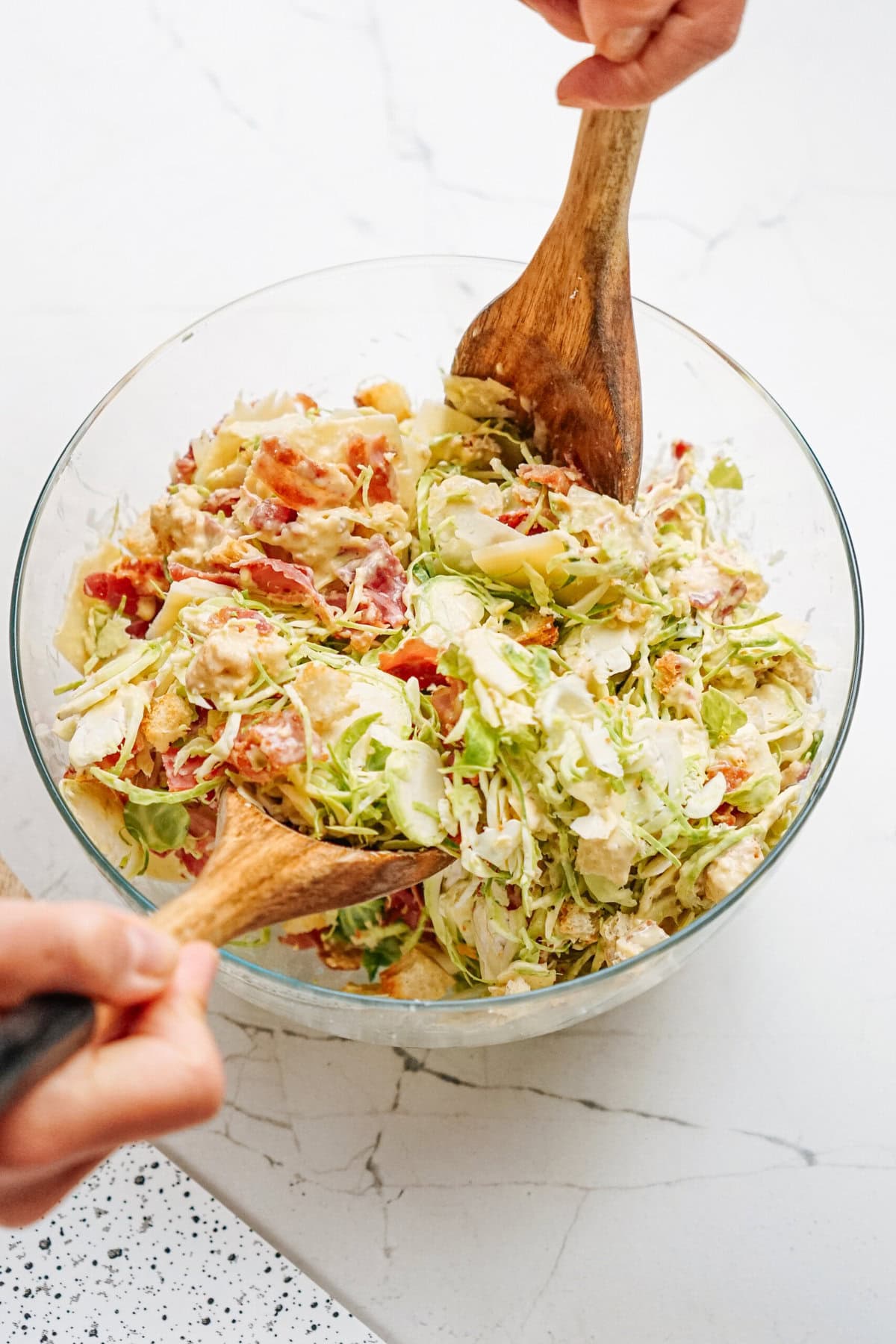 Hands using wooden spoons to mix a large glass bowl of salad with shredded lettuce, tomatoes, and other ingredients on a marbled countertop.