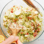 Hands mixing a salad with bacon and Brussels sprouts in a glass bowl using wooden utensils.