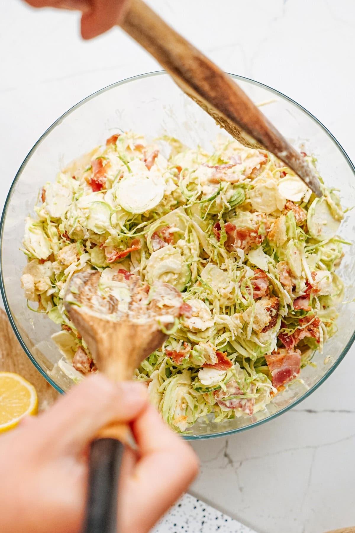 Hands mixing a salad with bacon and Brussels sprouts in a glass bowl using wooden utensils.