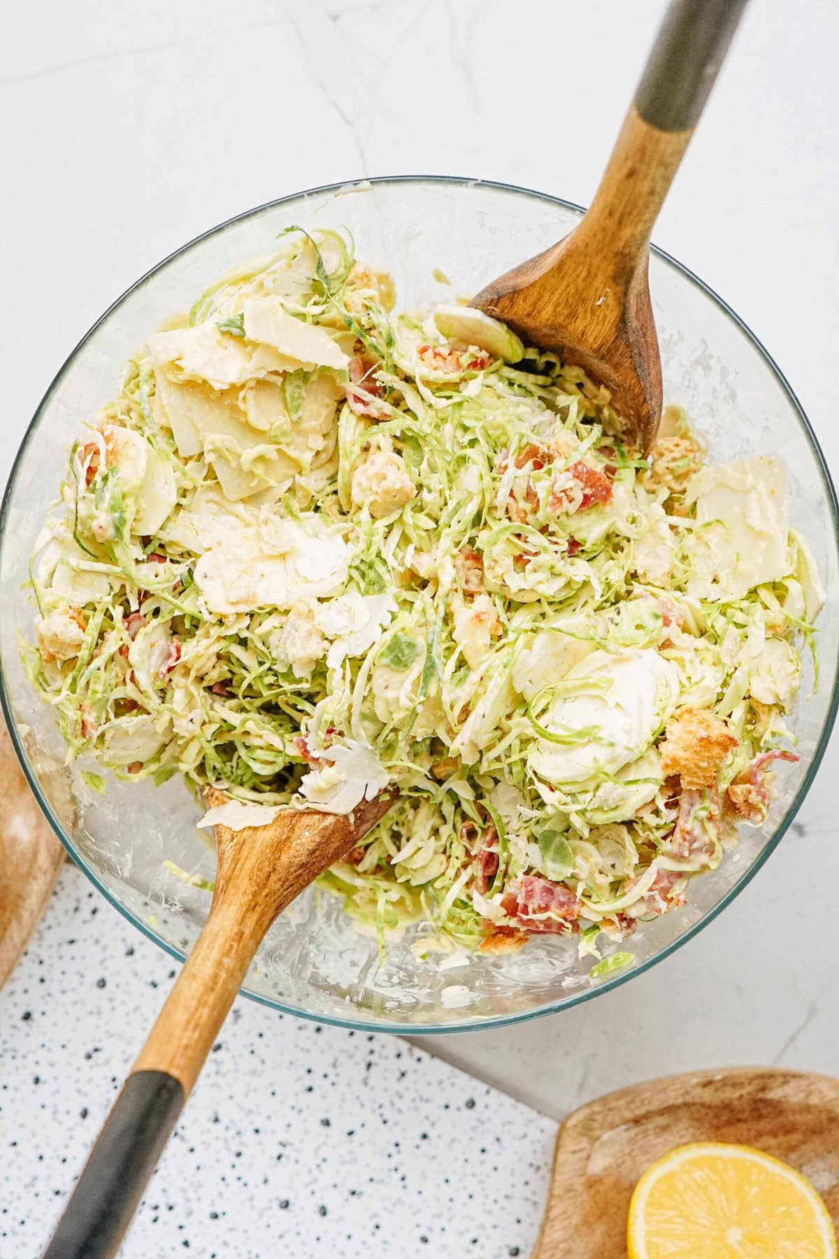 A glass bowl filled with a salad of shredded Brussels sprouts, tomatoes, parmesan cheese, and dressing. Two wooden serving utensils are in the bowl. A lemon half is on the side.