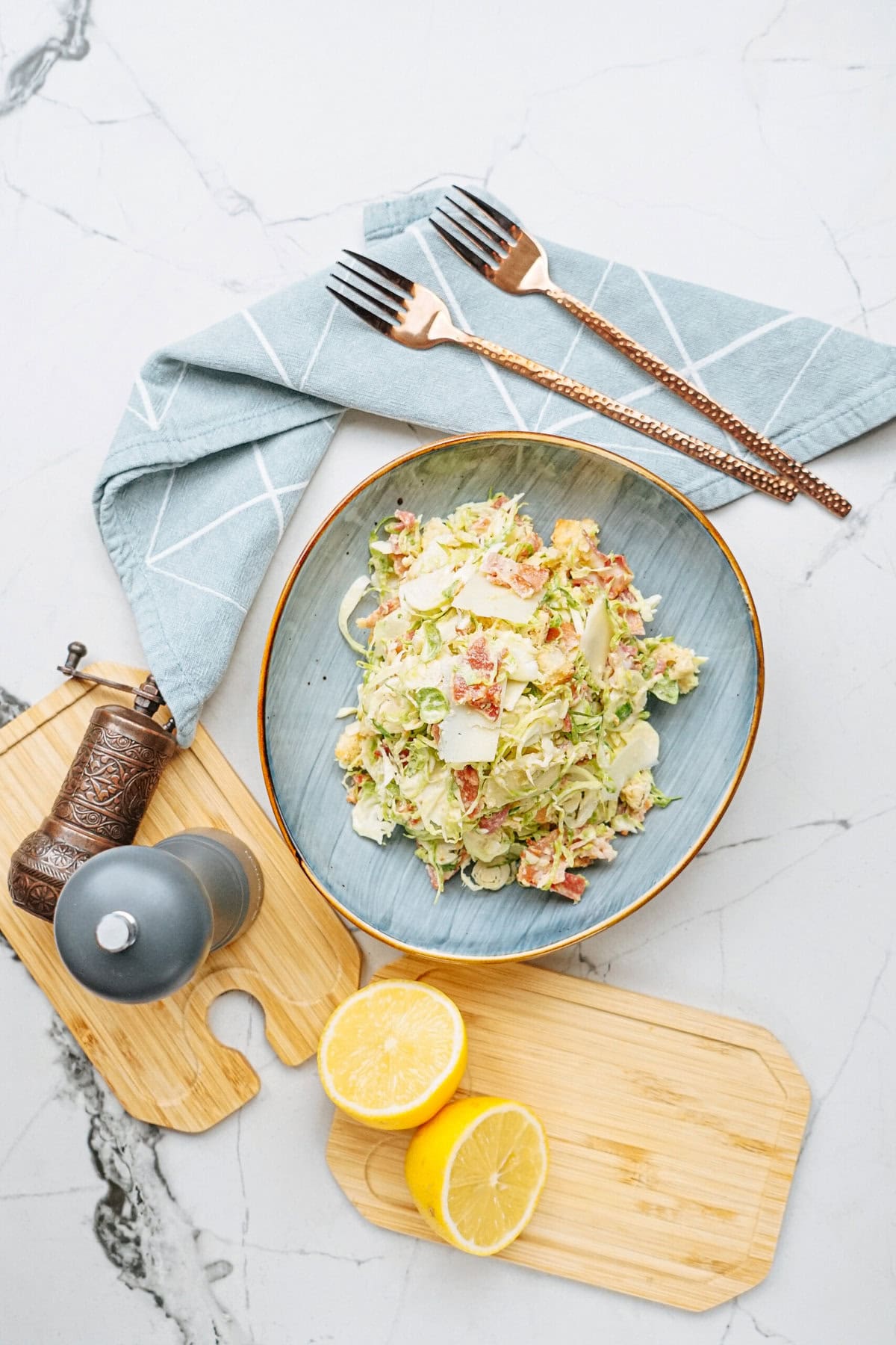 A bowl of salad with shredded lettuce and sliced toppings on a blue plate, accompanied by lemon halves, two forks on a napkin, and a pepper mill on a marble surface.