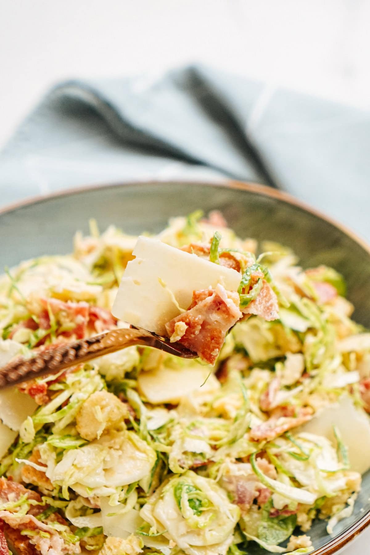 A fork holding a piece of cheese over a bowl of salad with shredded greens, bacon, and cheese. A folded cloth napkin is in the background.