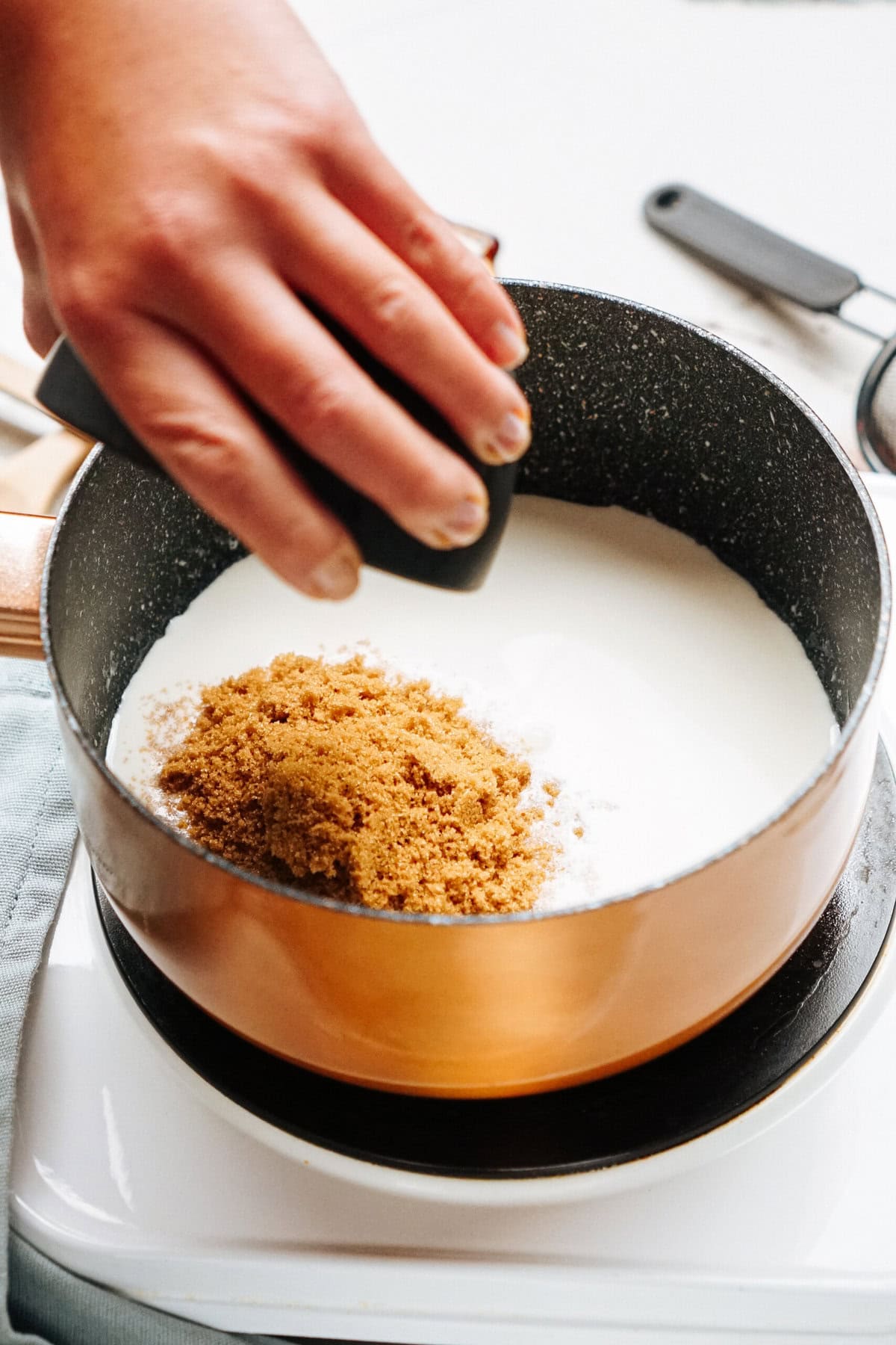 A hand sprinkling brown sugar into a pot of milk on a stove.