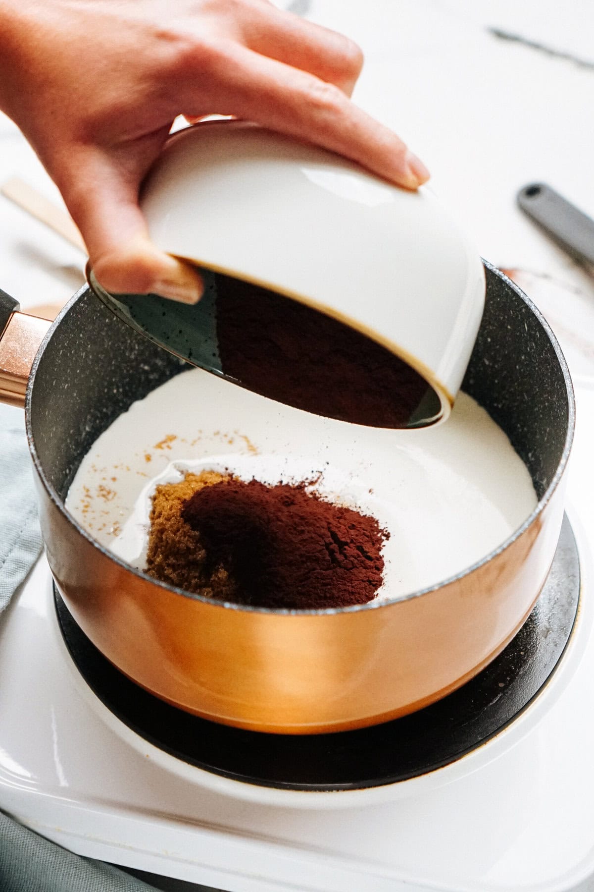 A hand pours cocoa powder from a white bowl into a saucepan containing a cream mixture on a stove.