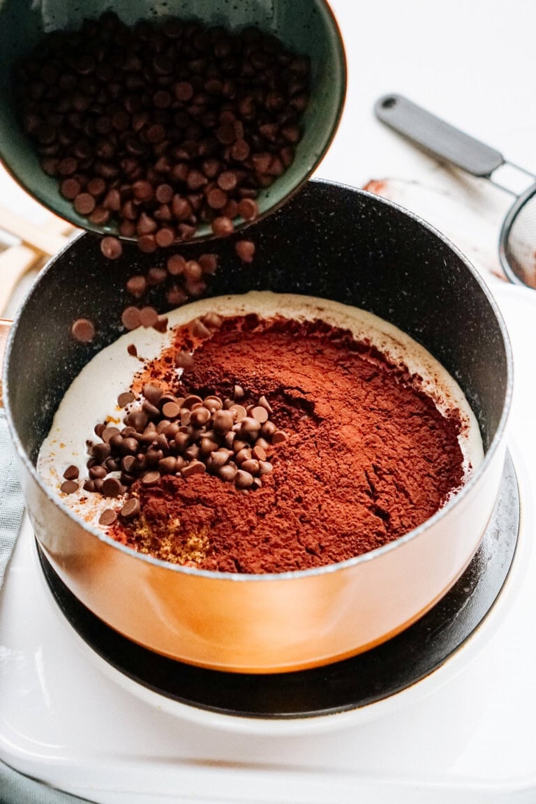 Chocolate chips and cocoa powder are being poured into a pot with cream, preparing to be mixed and melted.