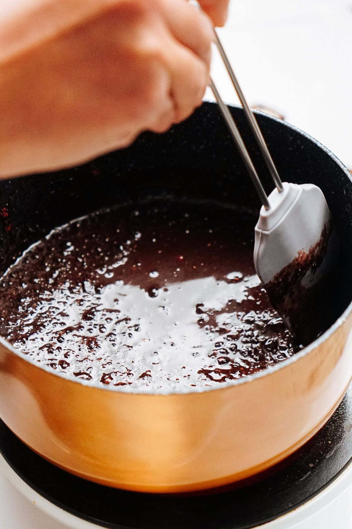 A hand stirring chocolate sauce in a copper pot on a stovetop.