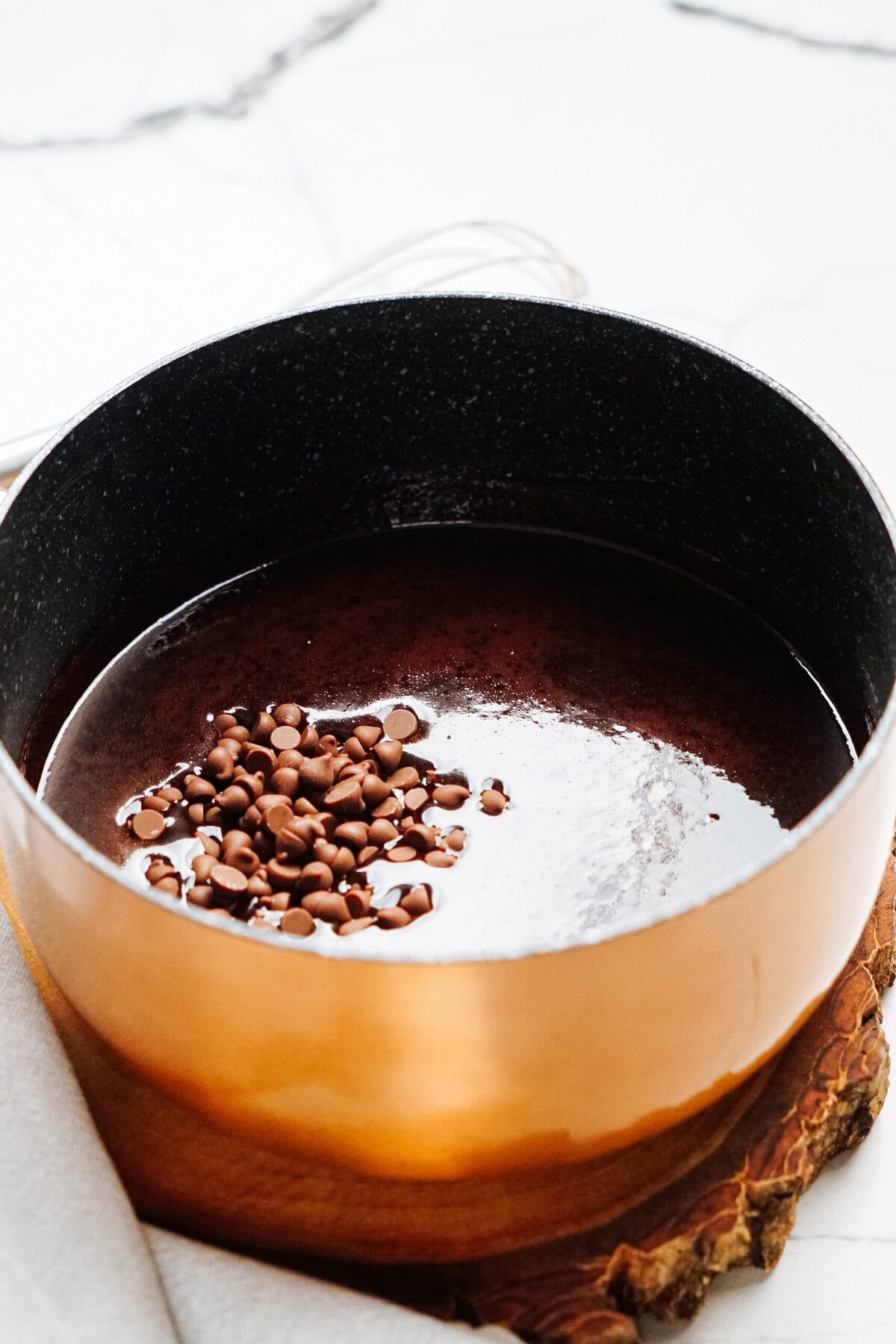 A copper pot filled with melted chocolate and topped with chocolate chips rests on a wooden board.