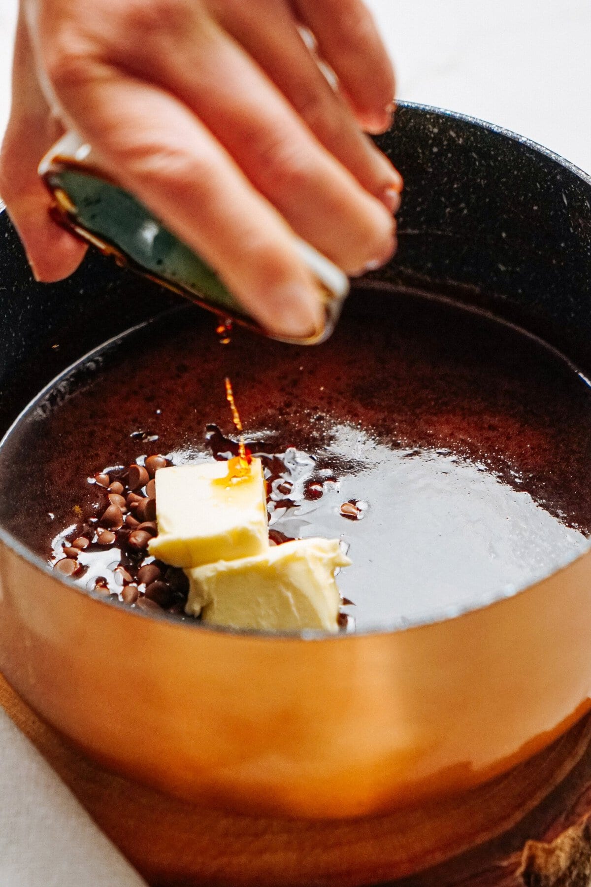 Close-up of a hand pouring a liquid into a copper saucepan containing chocolate chips, butter, and coffee on a stove.