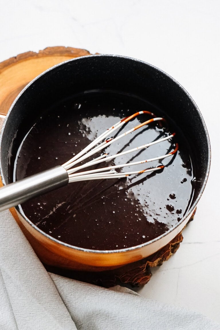 A whisk in a pot stirring dark liquid, likely chocolate sauce or syrup, on a wooden trivet with a cloth nearby.