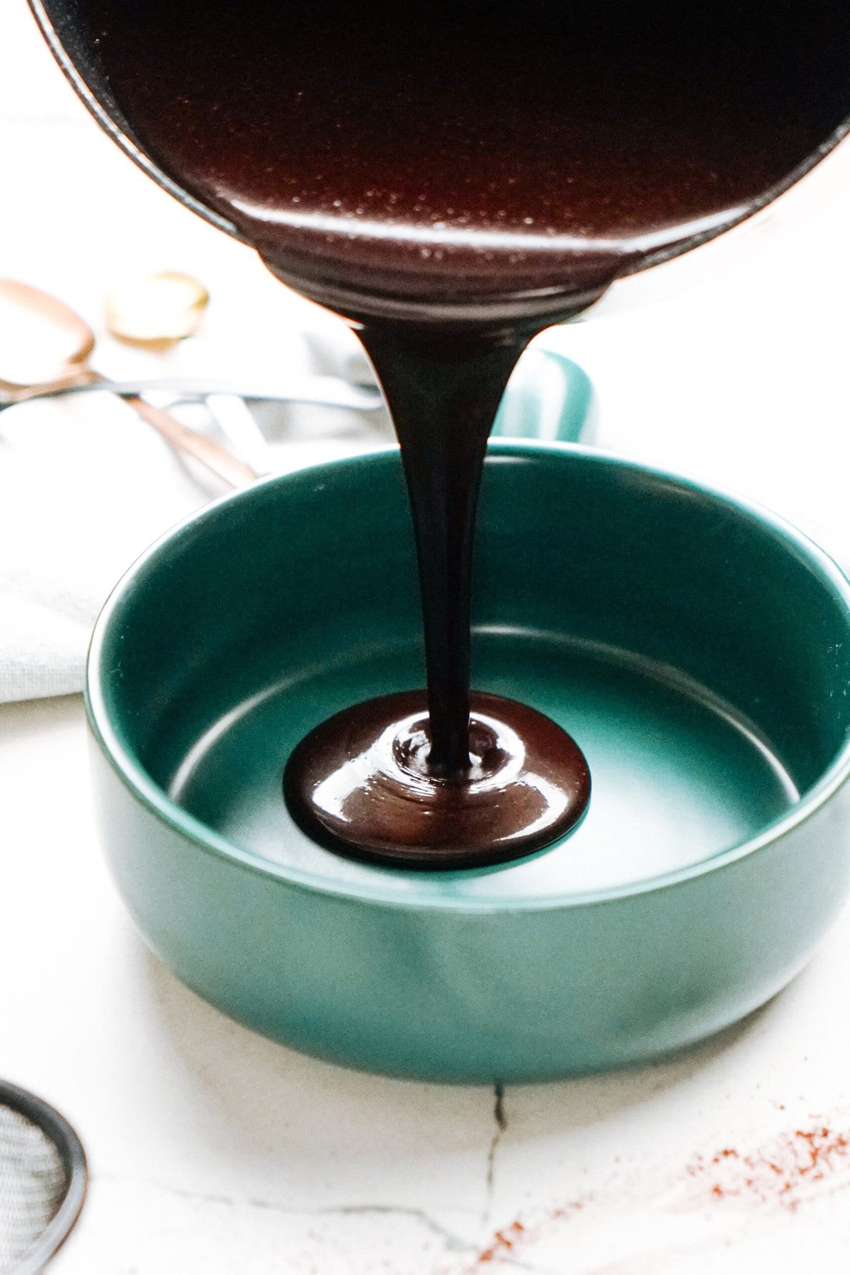 Chocolate sauce being poured into a teal bowl on a marble surface with kitchen utensils in the background.