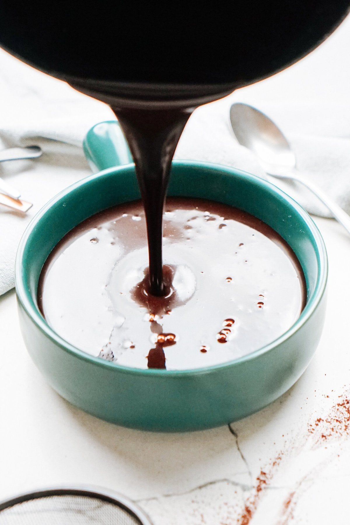 Pouring chocolate sauce into a teal bowl on a white surface with a spoon and whisk nearby.