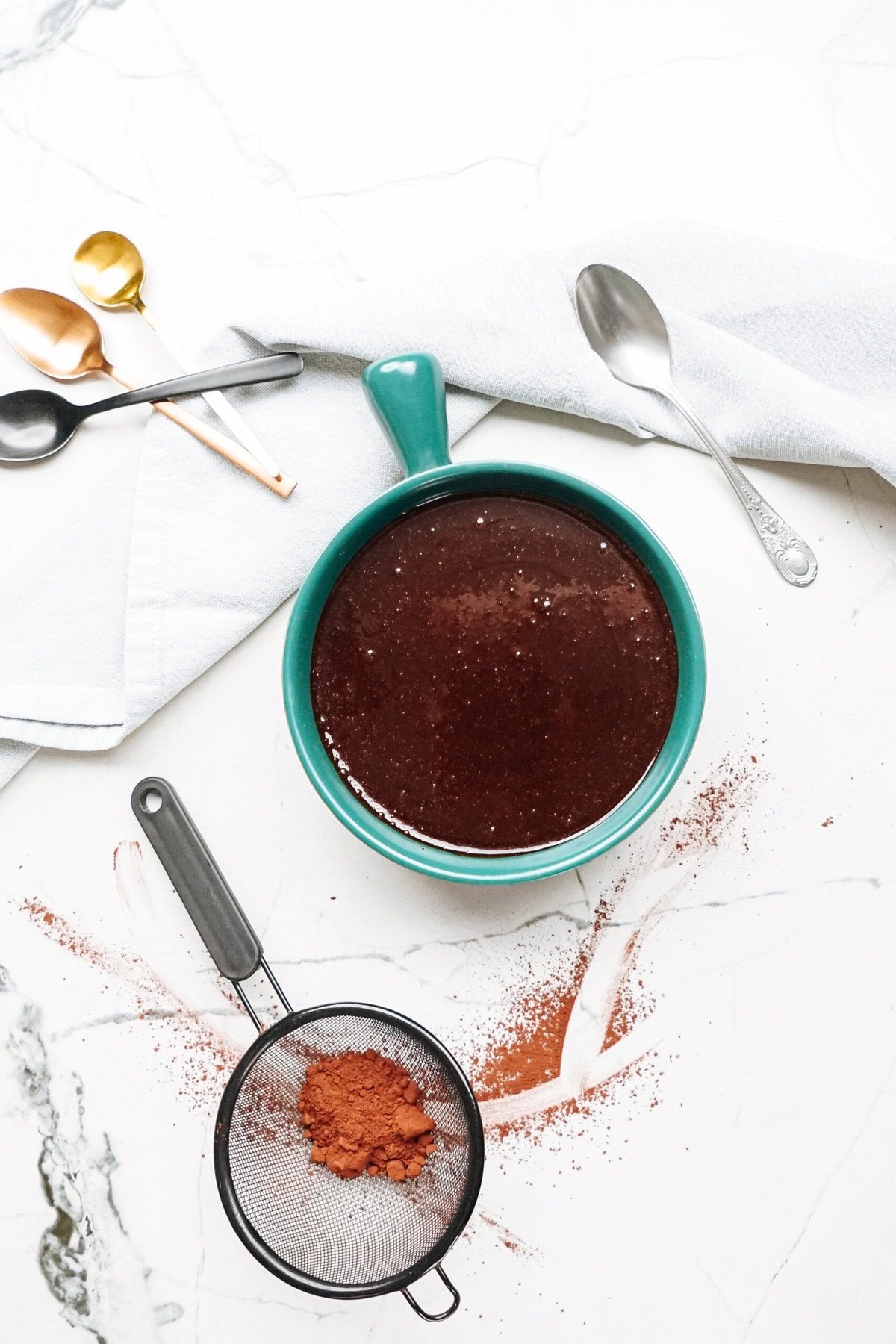 A green mug filled with hot fudge sauce, surrounded by a white towel, three spoons, a sifter with cocoa powder, and cocoa powder scattered on a white marble surface.