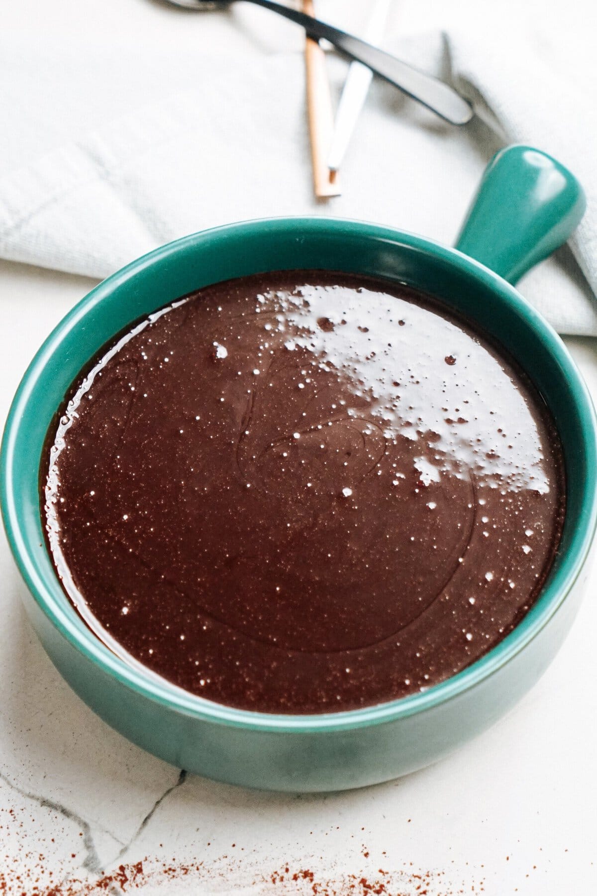 A teal mug filled with rich, dark hot fudge sauce sits on a white surface, with a light sprinkle of cocoa powder surrounding the rim.