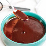 A spoon is being lifted from a bowl of dark brown liquid, dripping the liquid back into the bowl.