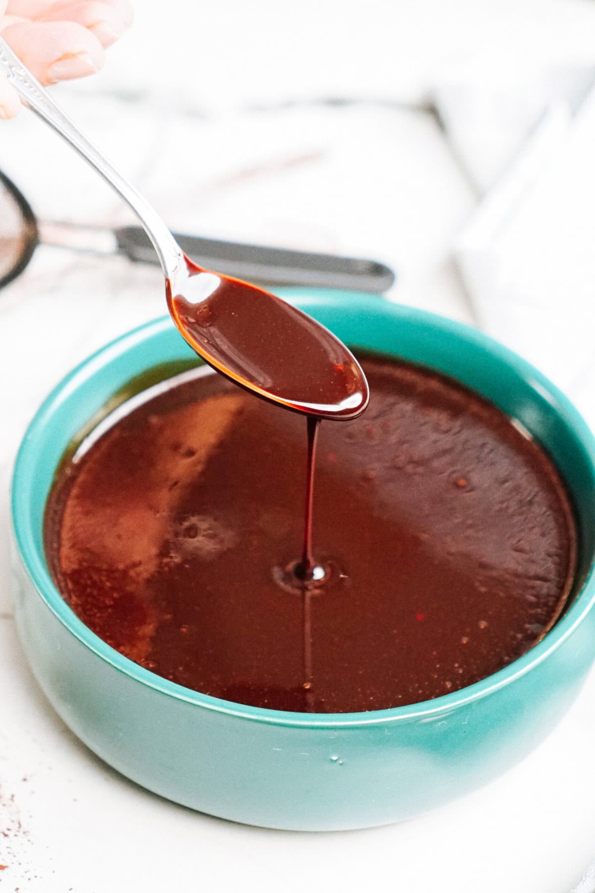 A spoon is being lifted from a bowl of dark brown liquid, dripping the liquid back into the bowl.