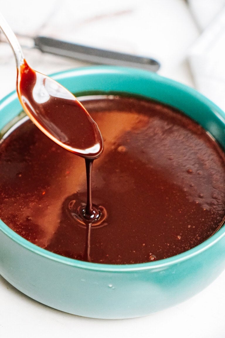 A spoon drizzles chocolate syrup into a round, teal bowl filled with chocolate sauce.