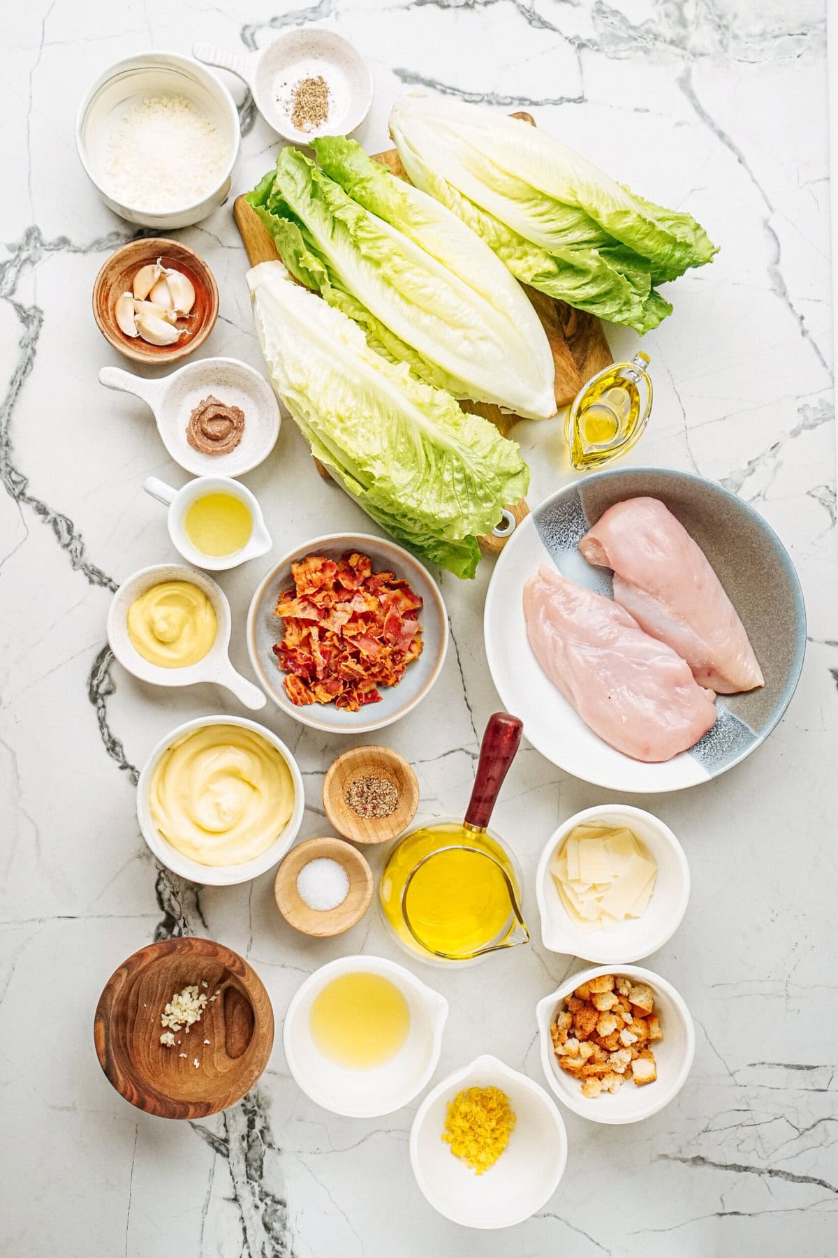 Ingredients for a salad are arranged on a marble surface, including romaine lettuce, chicken breasts, croutons, cheese, bacon, seasonings, and various dressings in bowls and containers.