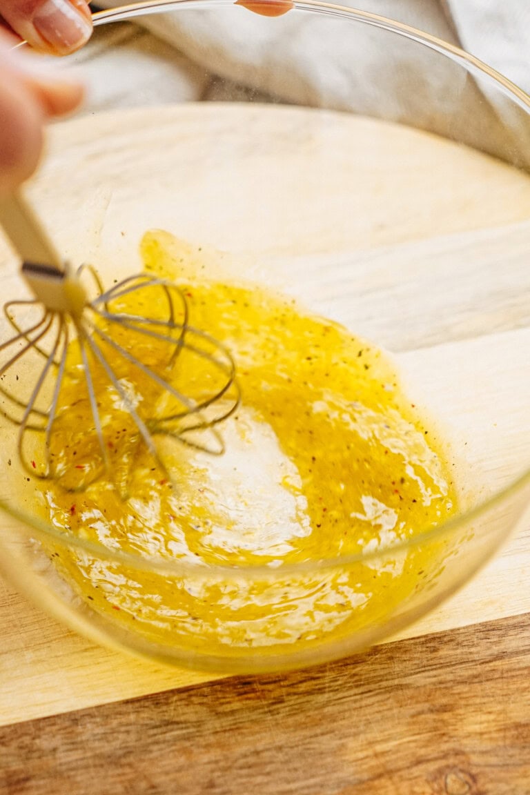 Close-up of a hand whisking yellow vinaigrette in a clear glass bowl on a wooden surface.