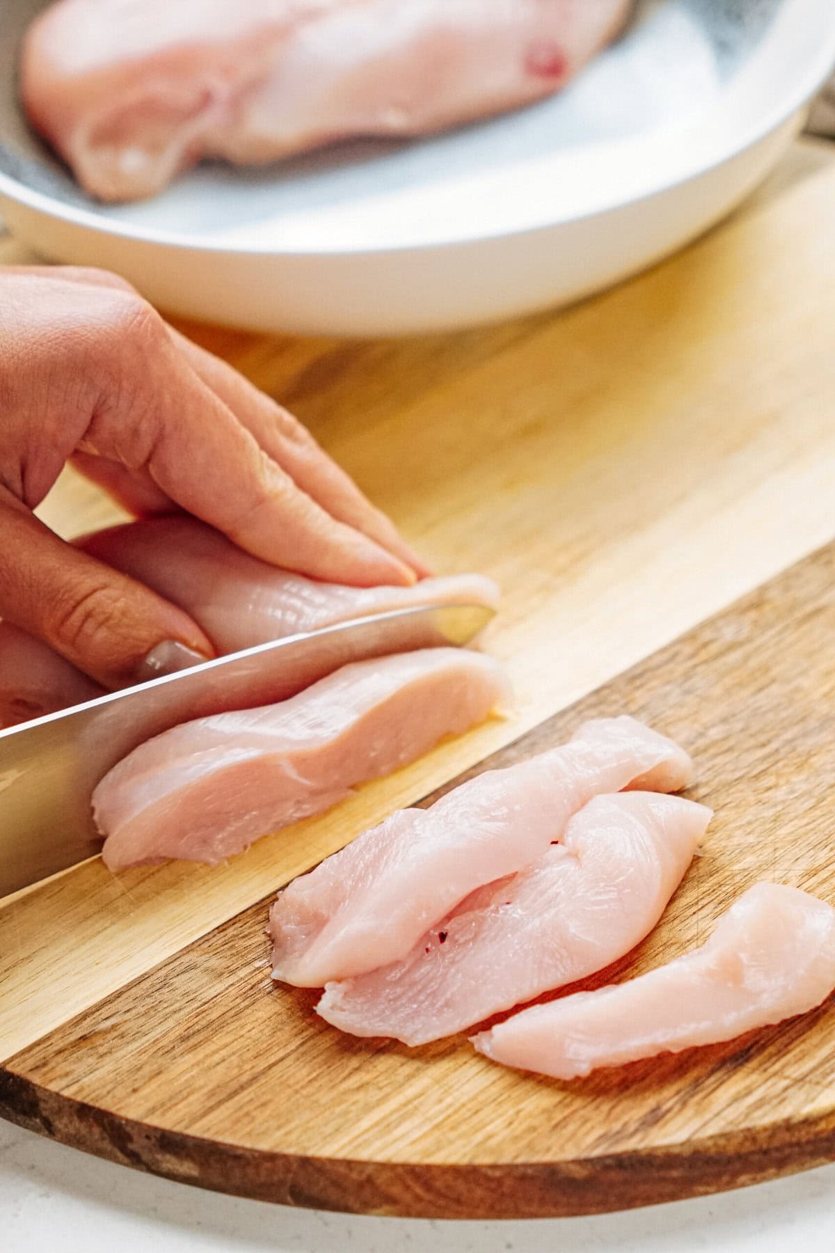 A person slicing raw chicken breast on a wooden cutting board with a knife.