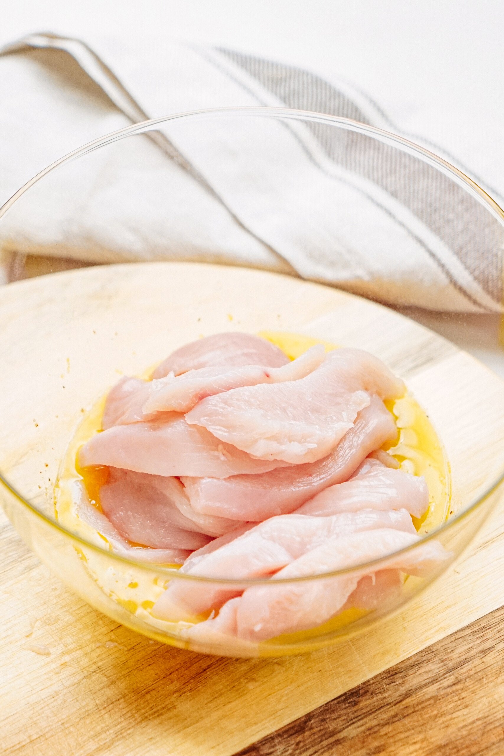 Raw chicken slices marinating in a glass bowl with a light-colored marinade on a wooden surface, with a cloth in the background.