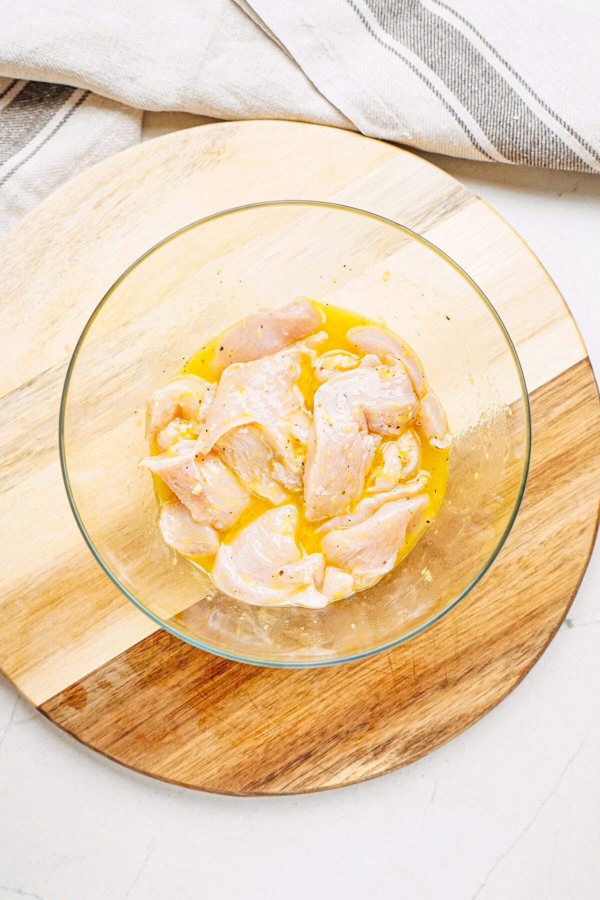 Raw chicken pieces marinating in a glass bowl with yellow liquid, placed on a wooden cutting board with a cloth nearby.