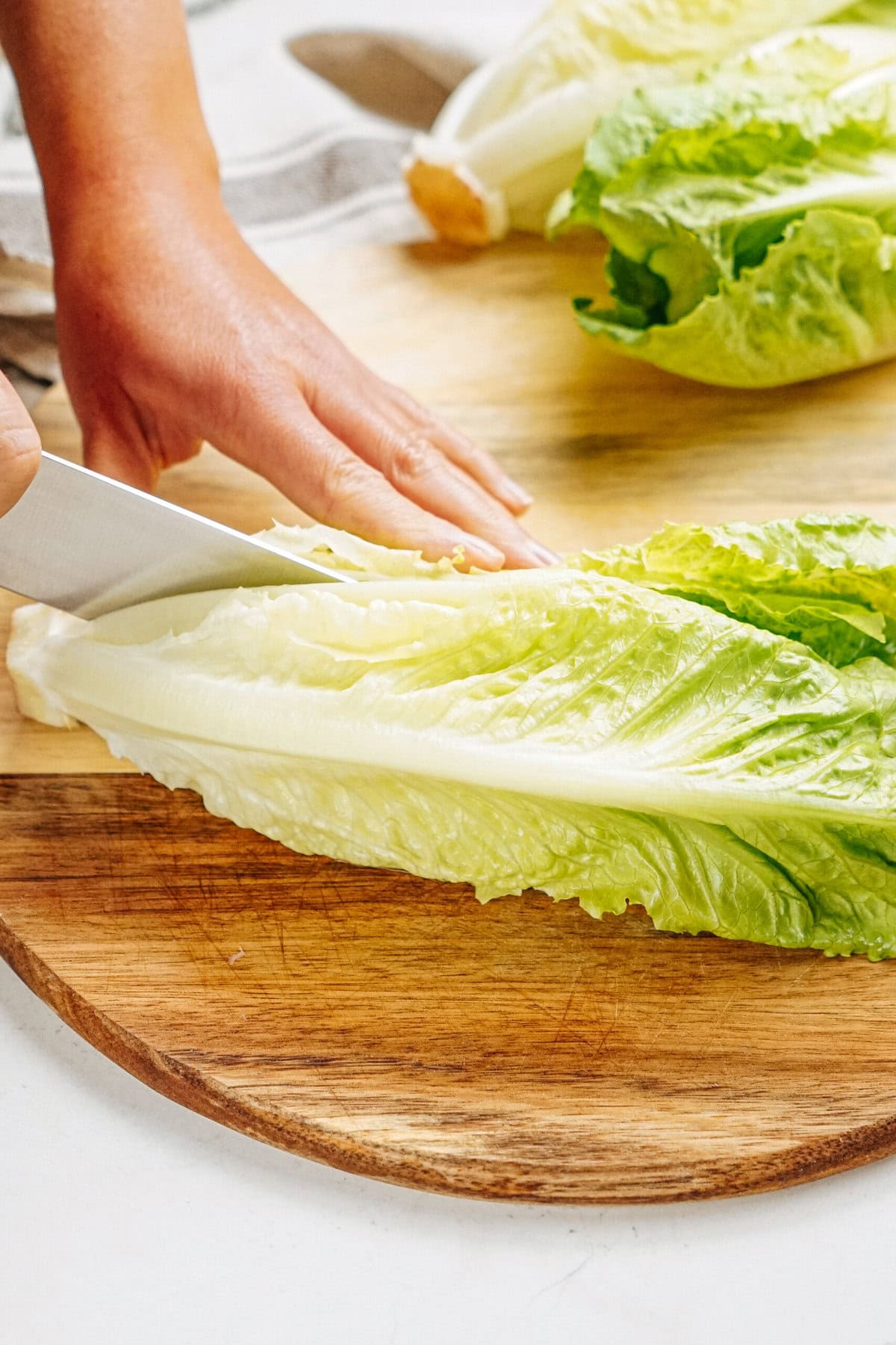 A person slices fresh romaine lettuce on a wooden cutting board.