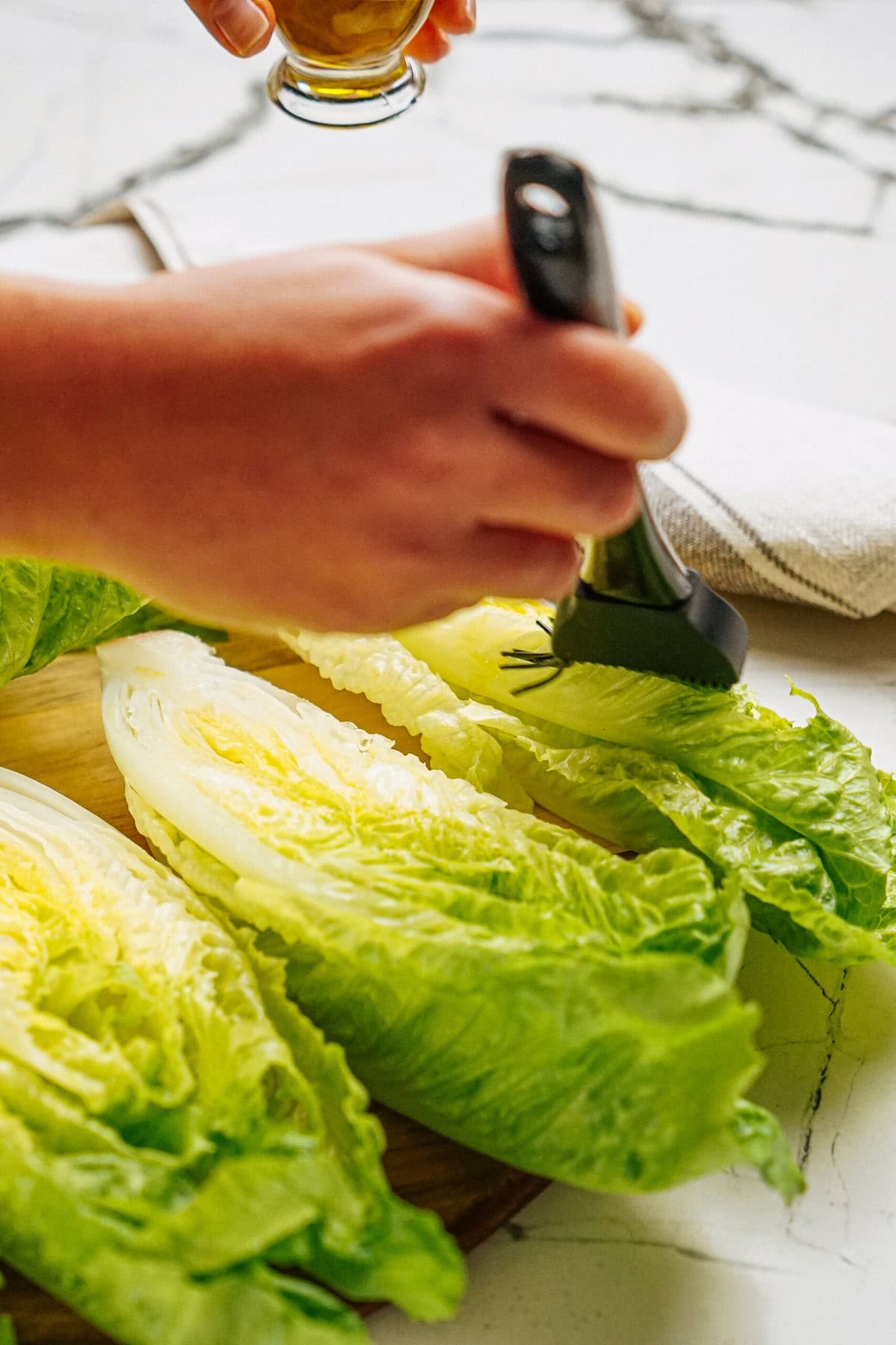 A person brushes dressing onto halved romaine lettuce on a wooden board placed on a marble countertop.