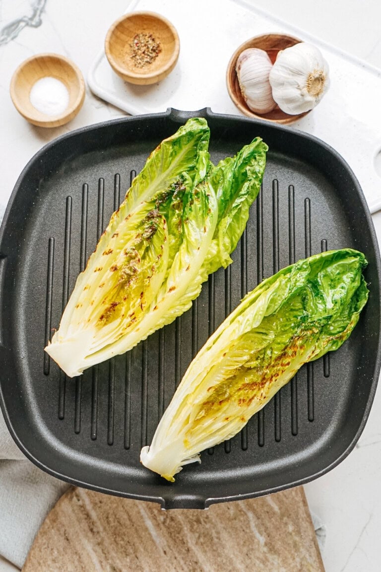 Two halves of romaine lettuce being grilled on a black grill pan, with garlic and small bowls of seasoning in the background.