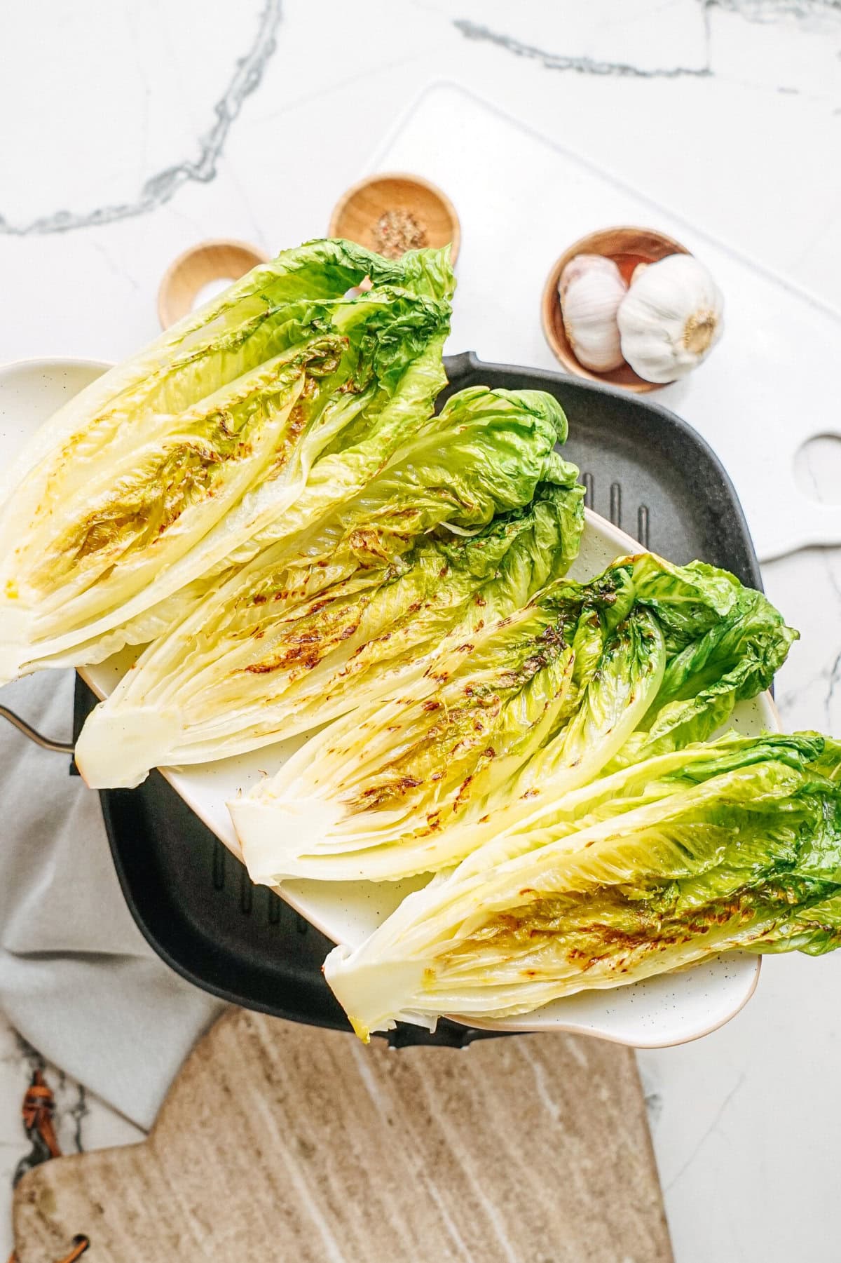 Grilled romaine lettuce halves on a plate, with garlic and spices in the background on a marble surface.